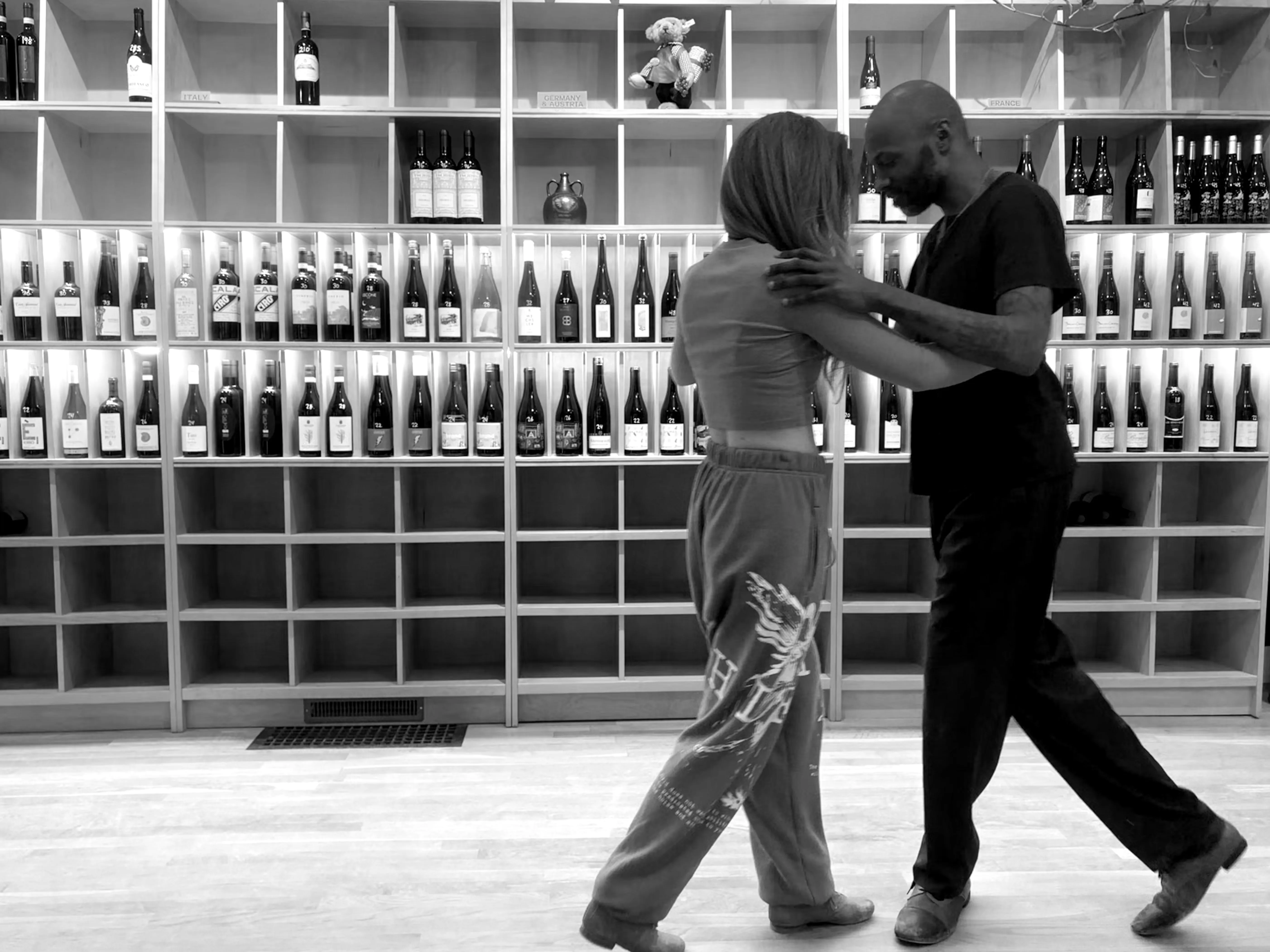 Two dancers taking a tango step in front of the wine shelves at Ritual Wines